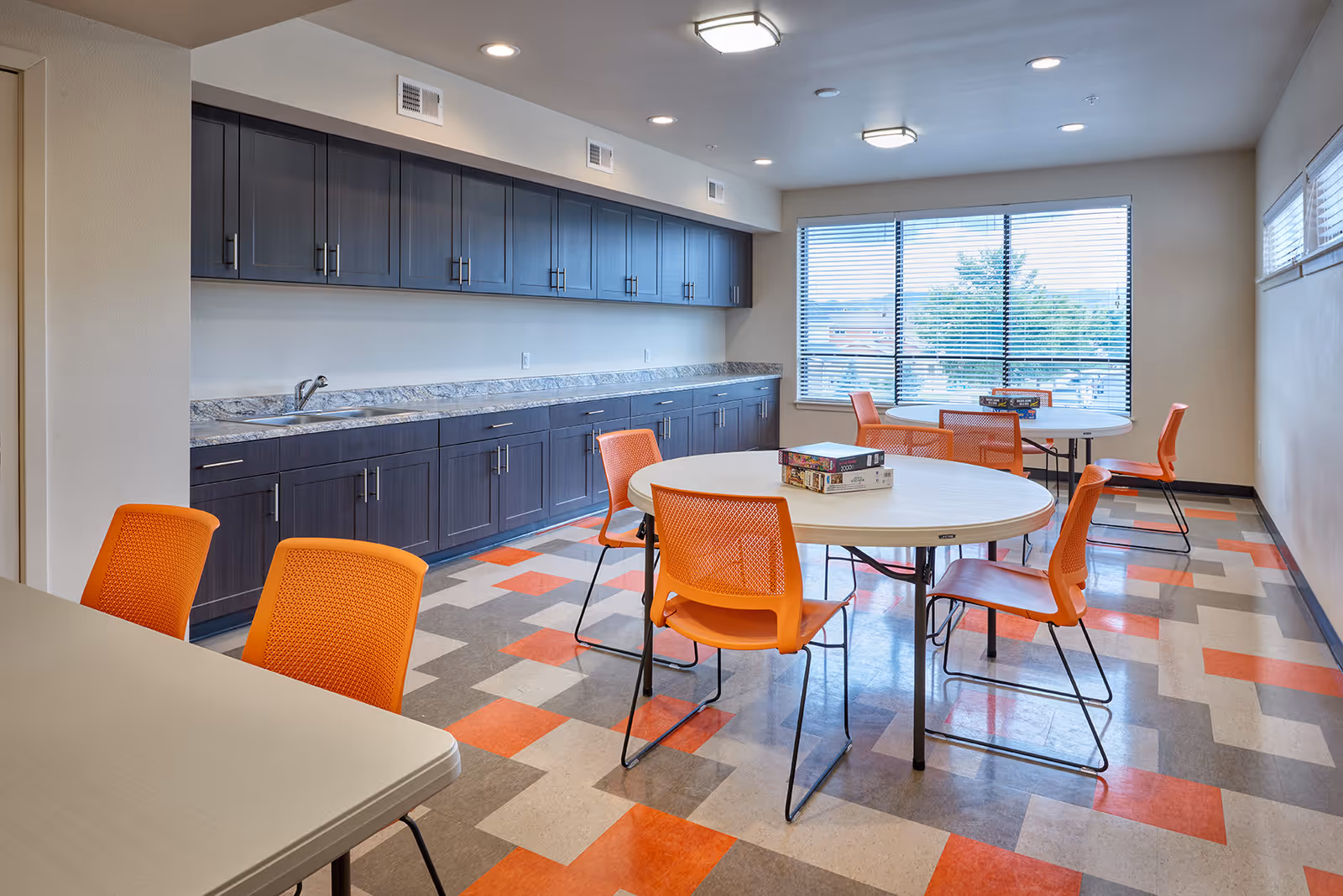 A bright activity room with large windows and blinds, featuring round tables with orange chairs. The floor has a colorful geometric pattern in orange, gray, and beige. Along one wall, there are dark wood cabinets with a marble countertop and a sink. Board games are placed on the tables.