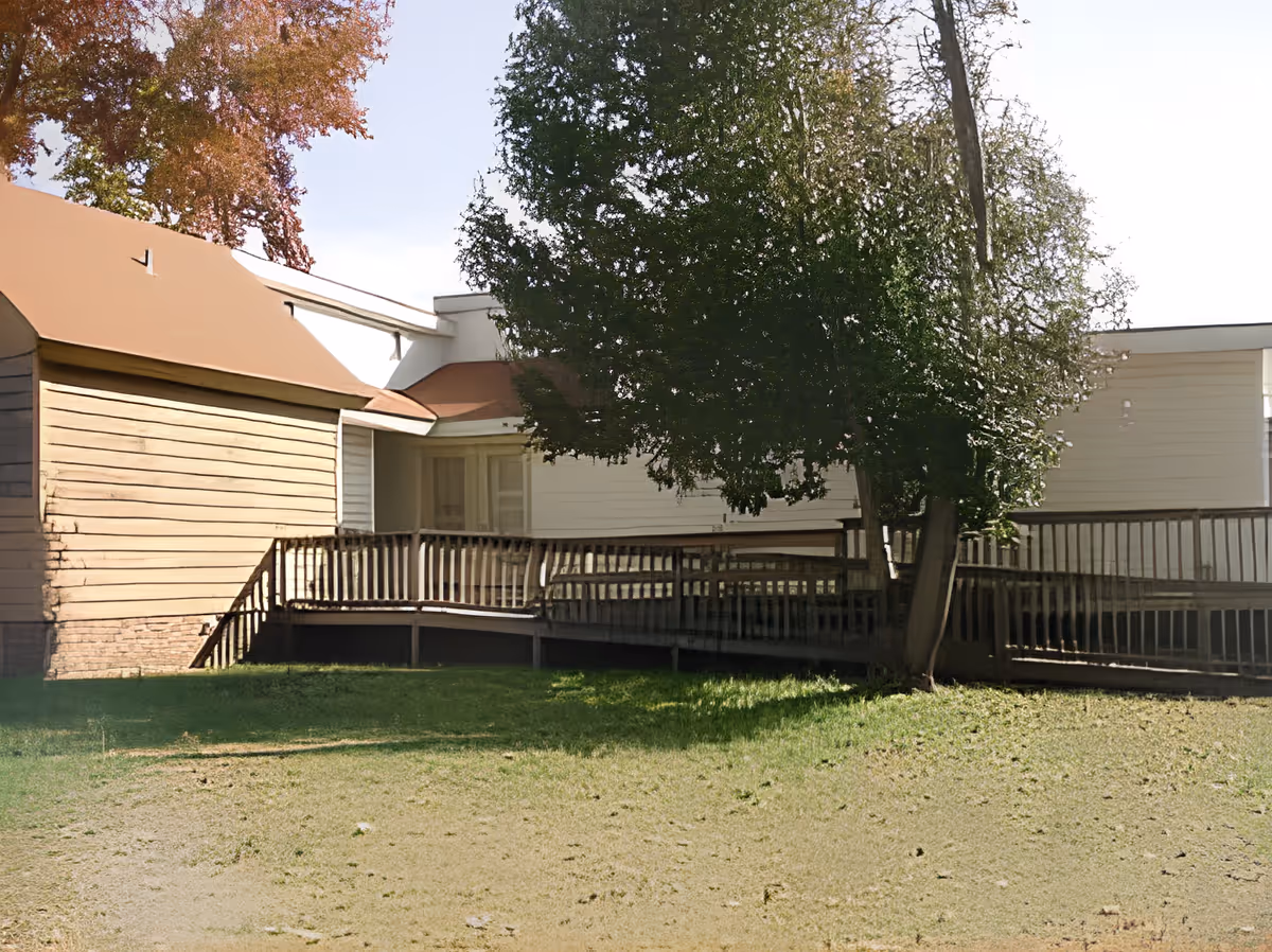 Exterior view of a single-story building with beige siding and a brown roof, featuring a wooden wheelchair-accessible ramp and railing. There is a large green tree in front of the building and a grassy lawn in the foreground.