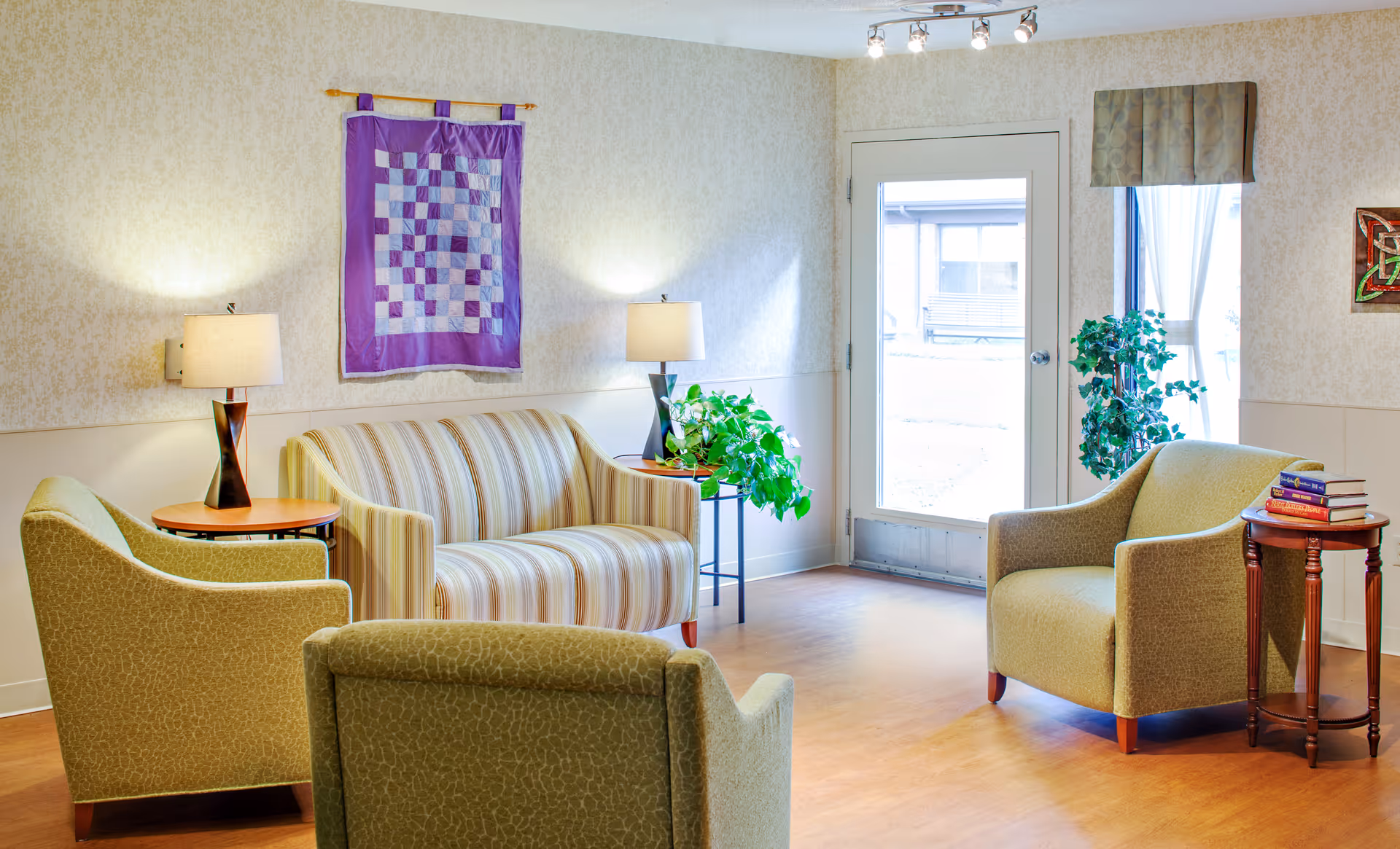 A cozy living room area with a striped loveseat, three green armchairs, two side tables with lamps, a small table with a green plant, a door with a window, and a window with curtains. A purple quilted wall hanging and a framed artwork decorate the walls.