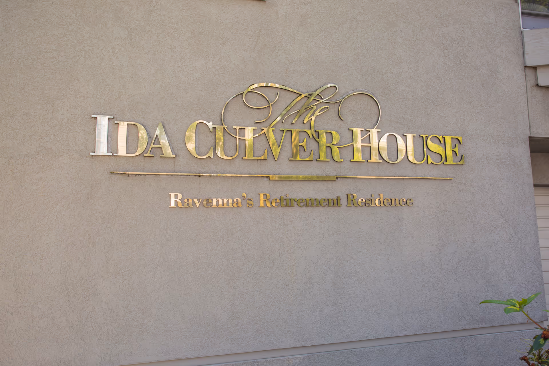 A beige exterior wall with a gold metallic sign that reads 'The Ida Culver House Ravenna's Retirement Residence'.