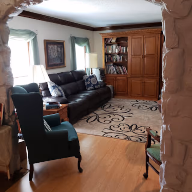 Living room with a dark leather sofa, two armchairs, a wooden cabinet/bookshelf and a patterned area rug viewed through a stone archway.