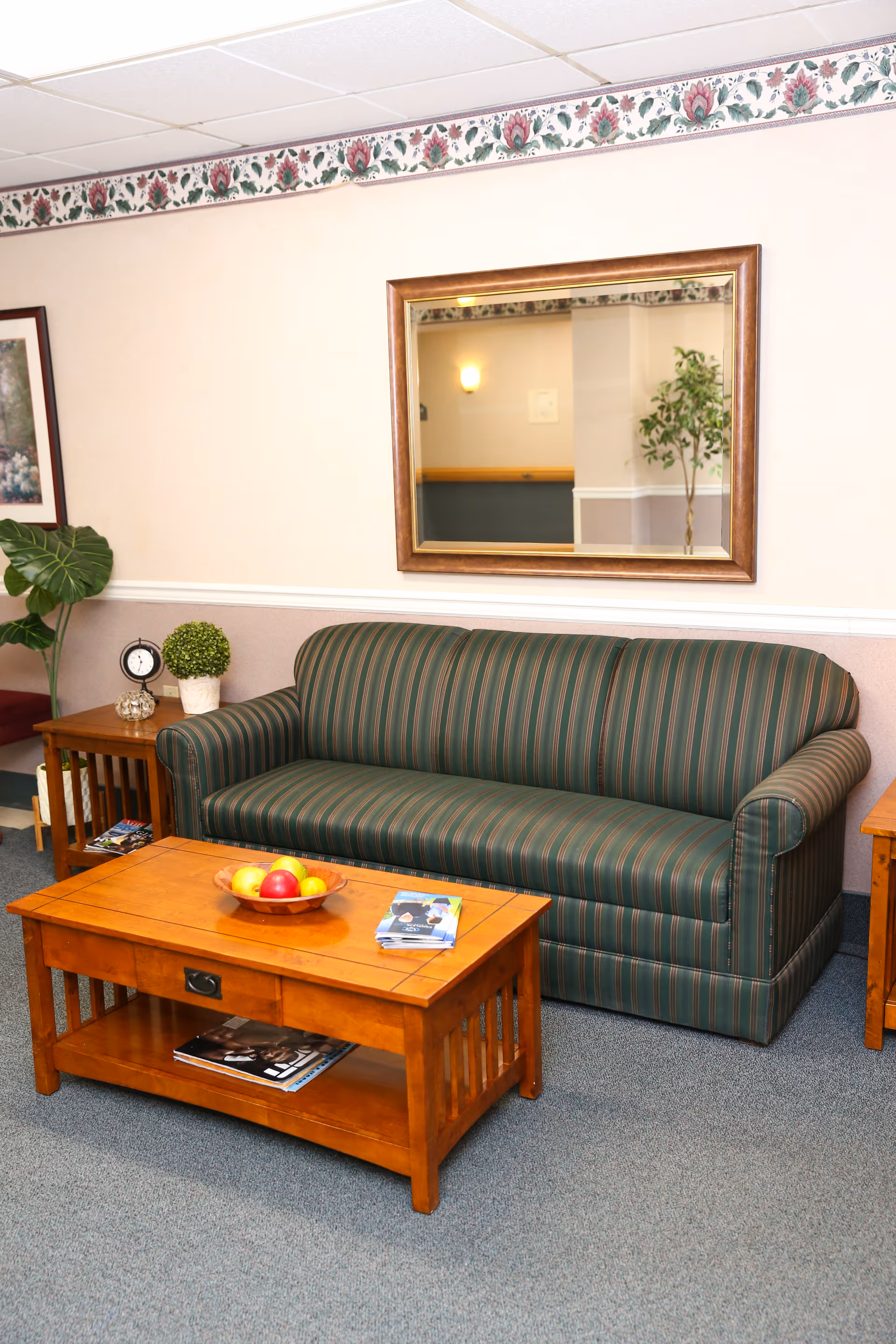 A cozy living room area with a green striped sofa, a wooden coffee table with a bowl of fruit and magazines, a side table with a small clock and plant, a large mirror on the wall, and a framed picture partially visible on the left.