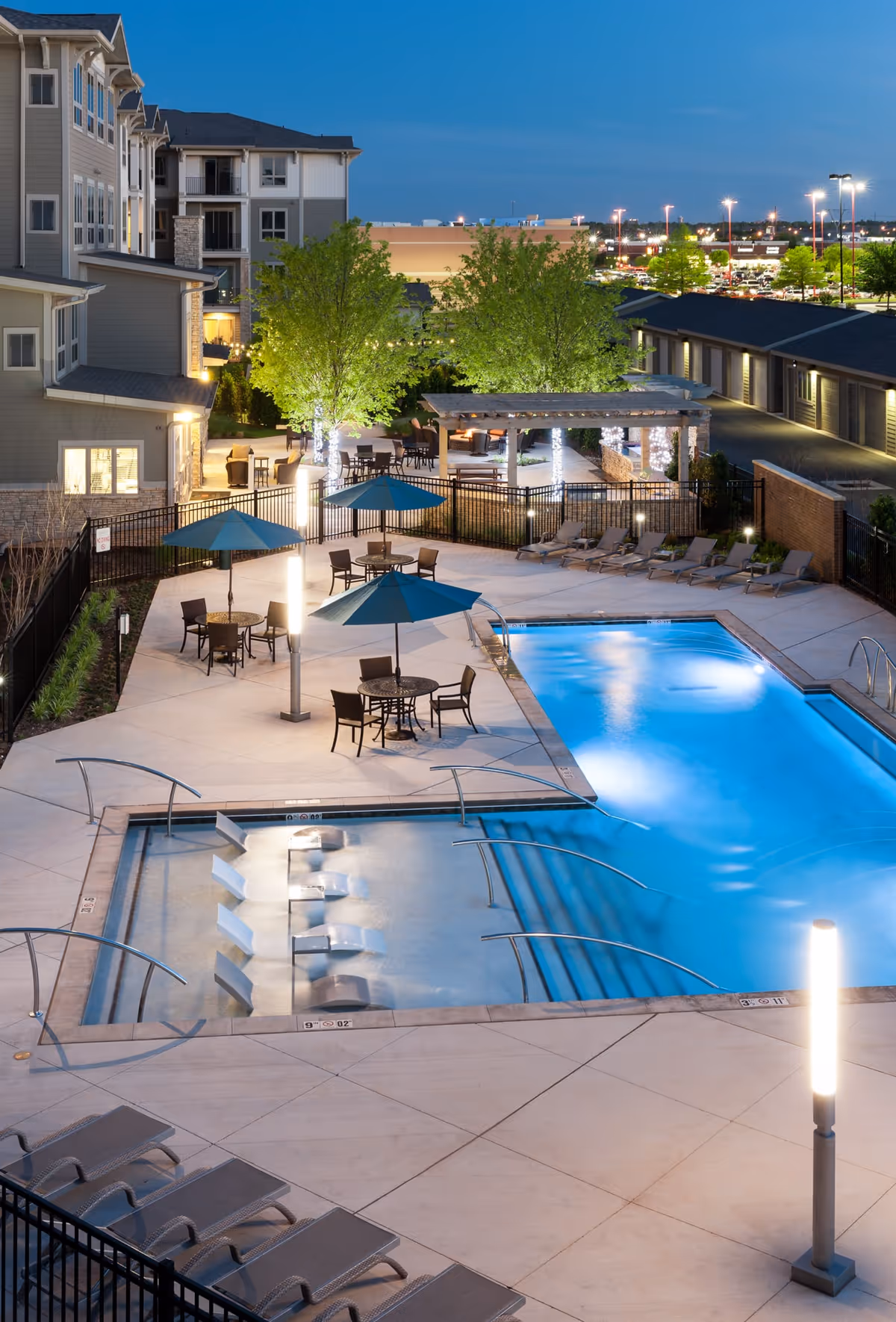 Outdoor swimming pool area at dusk with lounge chairs, tables with umbrellas, and a smaller pool with built-in seats. The area is surrounded by a fence and adjacent to a multi-story building with lit windows. Trees and a pergola with lights are visible in the background along with a parking lot and streetlights.
