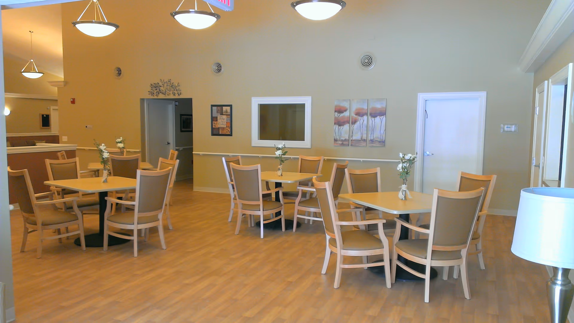 A dining area in a senior care facility with several square tables, each surrounded by four beige cushioned chairs. Each table has a small vase with white flowers. The room has light wood flooring, beige walls, and ceiling lights. There are two doors and some wall decorations including a framed picture and a three-panel artwork of trees.