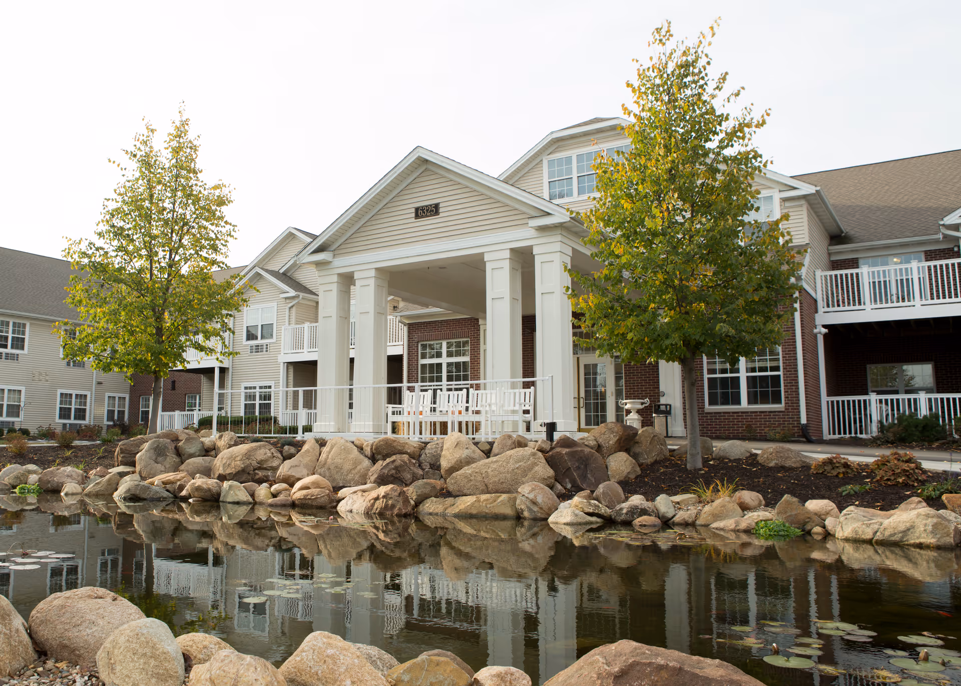 Exterior view of a senior living facility named The Keystones of Cedar Rapids, featuring a large building with white columns and a covered entrance. There are two trees with green leaves and a pond with rocks in the foreground reflecting the building and trees.