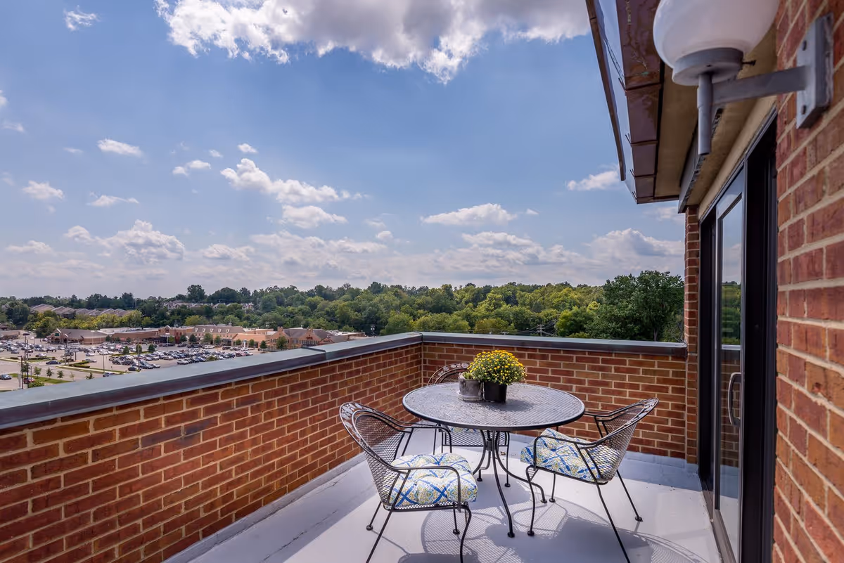 A rooftop balcony with a round metal table and three metal chairs with patterned cushions. The balcony has brick walls and overlooks a parking lot, buildings, and a green tree line under a partly cloudy blue sky.