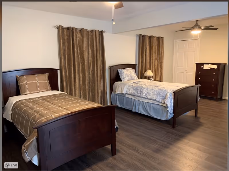 A senior care facility bedroom with two single beds, each with wooden headboards and footboards. One bed has a brown checkered comforter and pillow, while the other has a blue and white floral comforter and pillow. There are brown curtains on the windows, a wooden dresser with drawers, a small lamp on a nightstand, and ceiling fans with lights. The room has wood flooring and white walls.