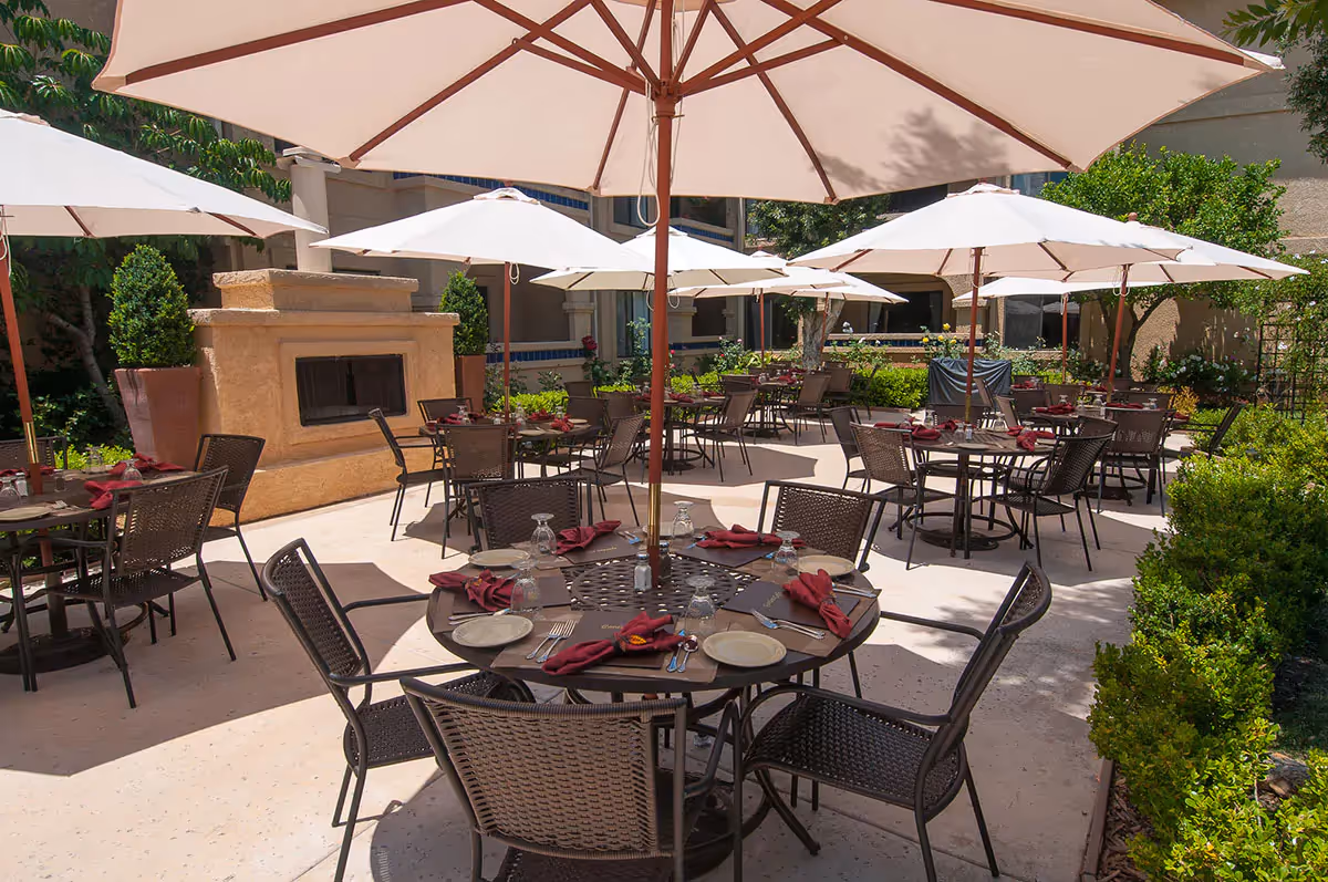 Outdoor patio dining area with multiple round tables set with plates, glasses, and burgundy napkins. Each table is shaded by large white umbrellas. There is a stone fireplace and potted plants along the perimeter, with greenery and bushes surrounding the patio.