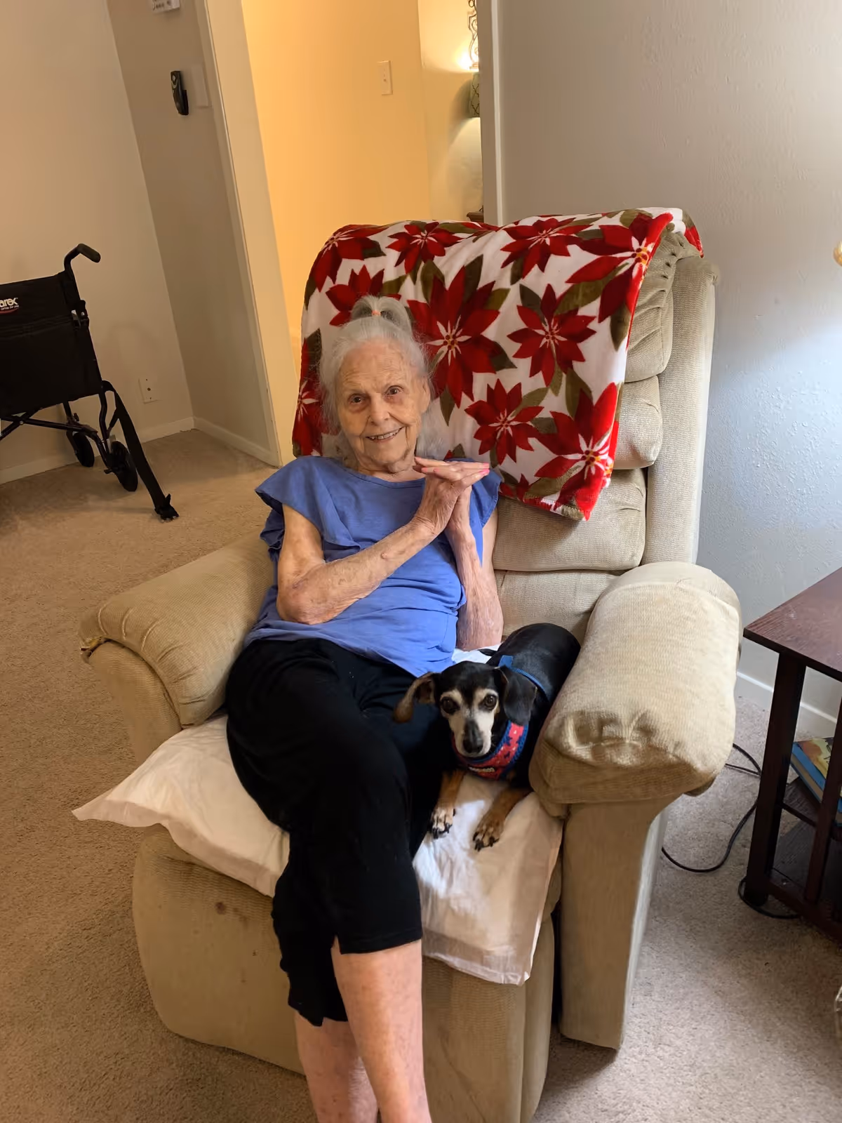 An elderly woman sitting in a beige recliner chair with a red and white floral blanket draped over the back. She is smiling and wearing a blue shirt and black pants. A small black and brown dog with a red collar is sitting on her lap. In the background, there is a black walker and a doorway leading to another room.
