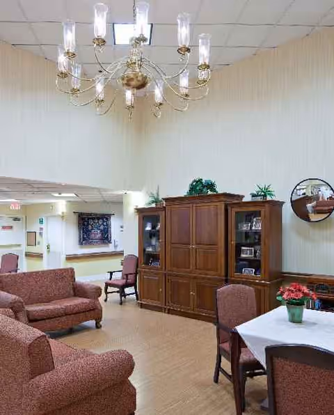 Lounge area with maroon sofas, wooden cabinet, chandelier, and dining table in a senior living facility.