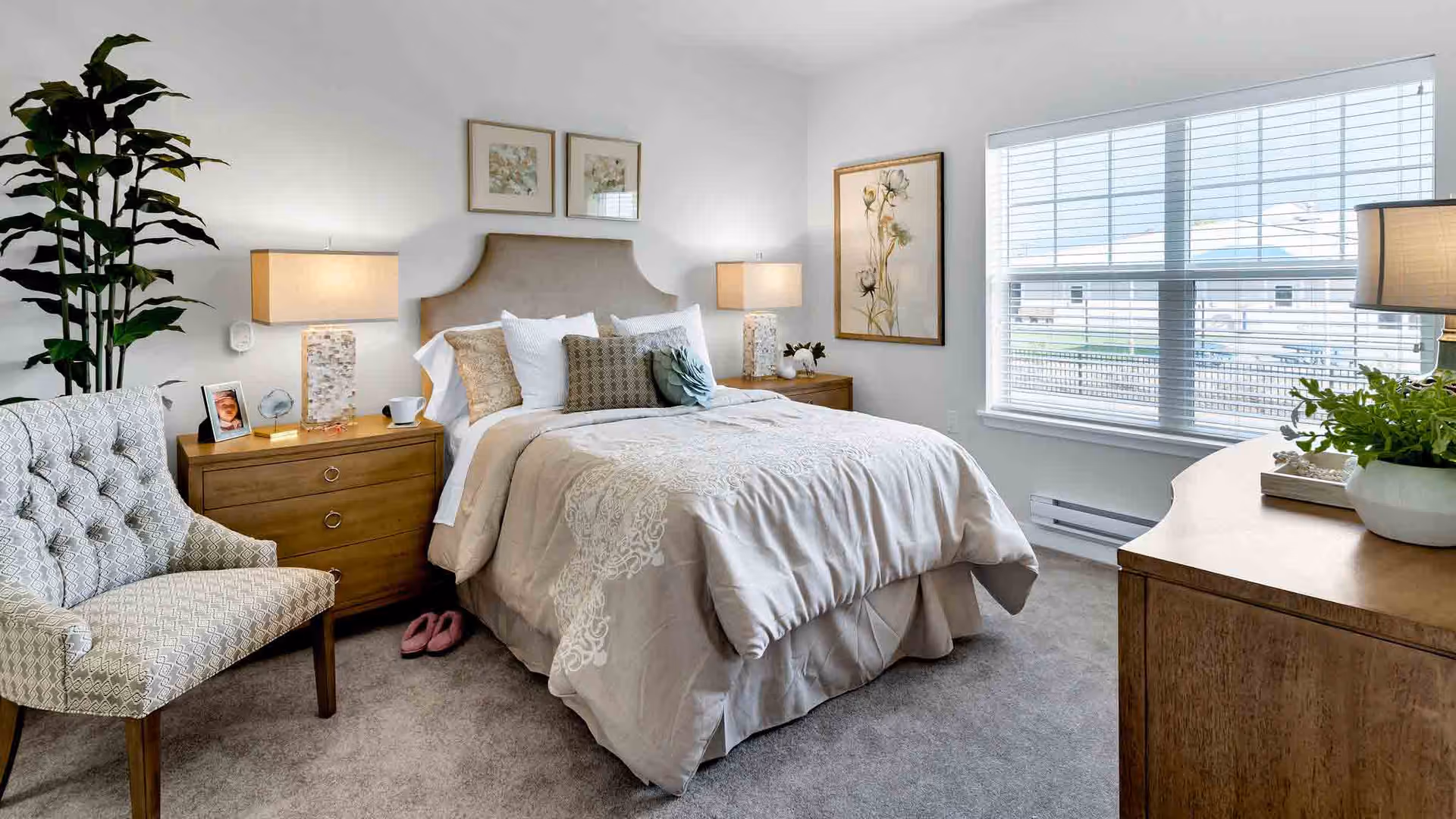 A cozy bedroom with a neatly made bed featuring beige and white bedding, two wooden nightstands with lamps on each side, a patterned armchair, a large window with blinds, a wooden dresser with a plant on top, and framed artwork on the walls.