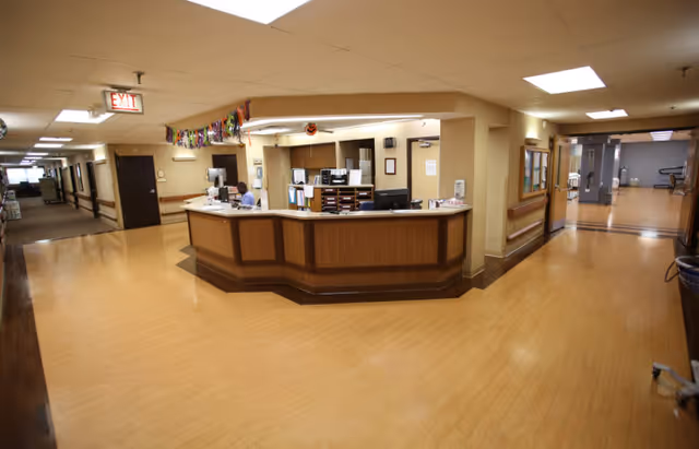 A central reception/nurses' station in a wide, well-lit senior living hallway with wood floors and ceiling lights.