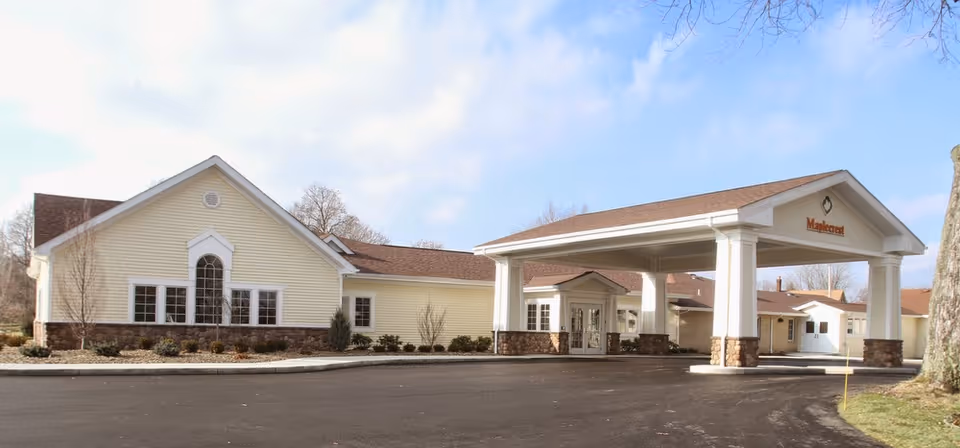Exterior view of Maplecrest Nursing Home showing a single-story building with beige siding, a covered entrance supported by white columns with stone bases, and a paved driveway in front. The sky is partly cloudy and there are some leafless trees in the background.