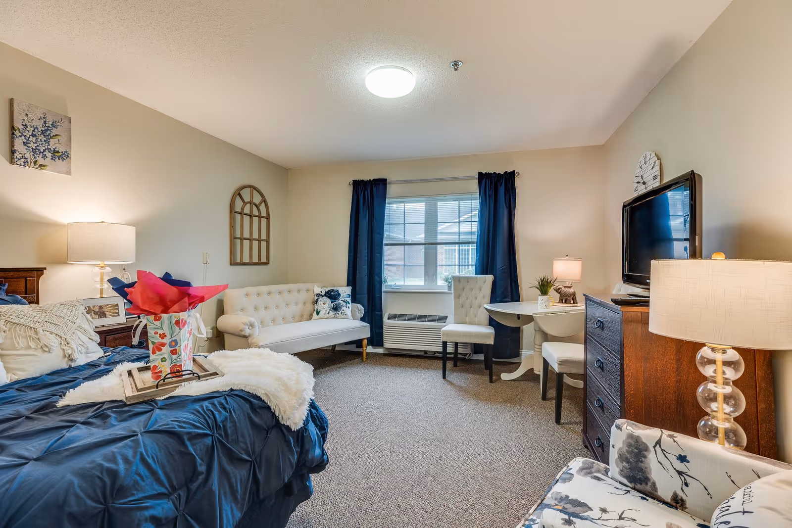 A cozy bedroom in an assisted living facility featuring a bed with a dark blue comforter and decorative pillows, a small white tufted sofa, a window with dark blue curtains, a small round table with two chairs, a wooden dresser with a flat-screen TV on top, and multiple lamps providing warm lighting.