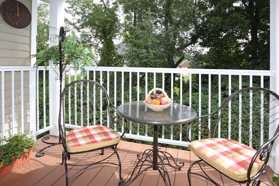 Outdoor patio area with two metal chairs with red and beige plaid cushions and a round glass-top table with a basket of fruit on it. The patio has a white railing and is surrounded by green trees.