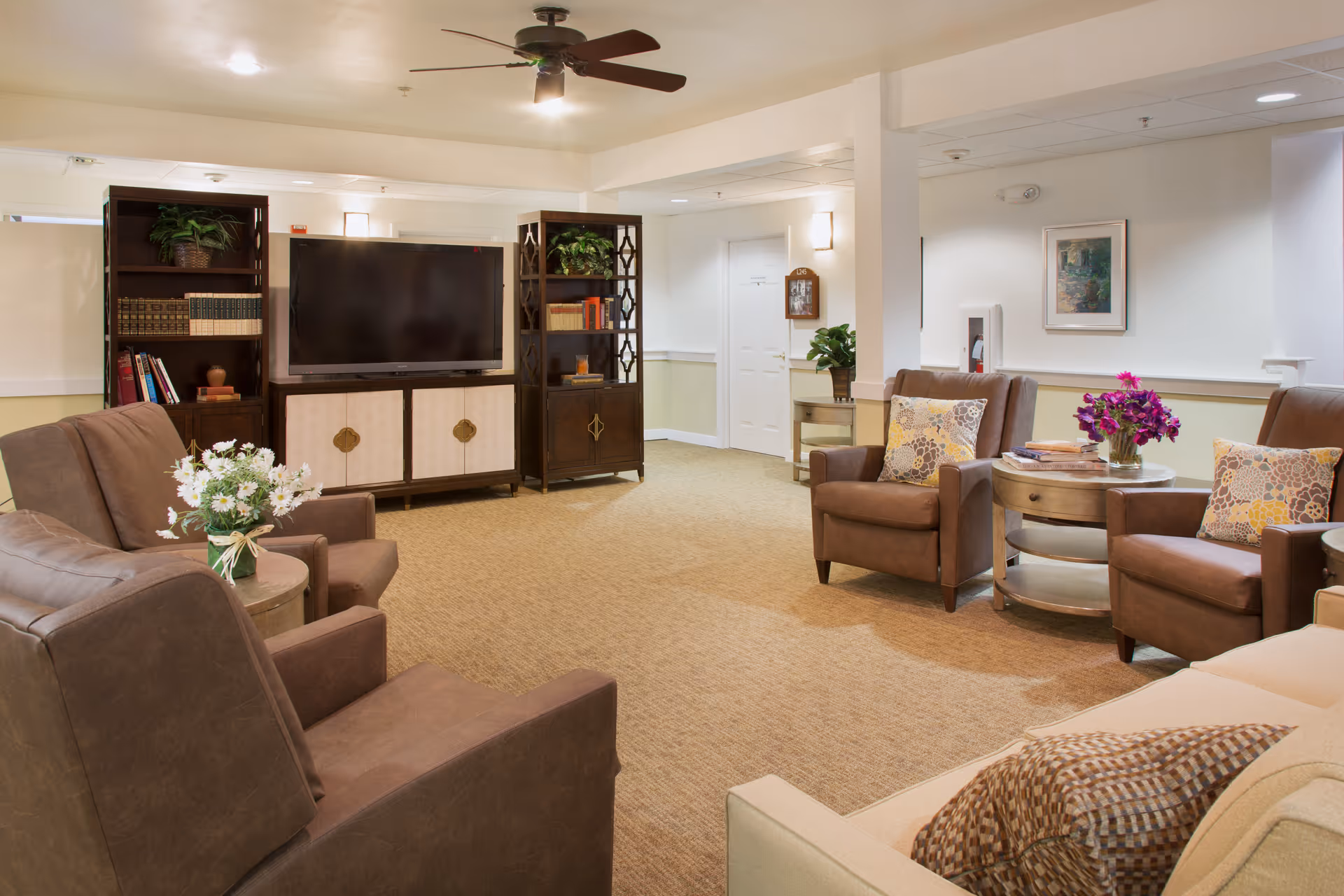 A cozy living room area with brown leather armchairs and a beige sofa arranged around small round tables with flower vases. A large flat-screen TV is mounted on a wooden entertainment center with bookshelves on either side. The room has beige carpeting, white walls, a ceiling fan, and soft lighting.
