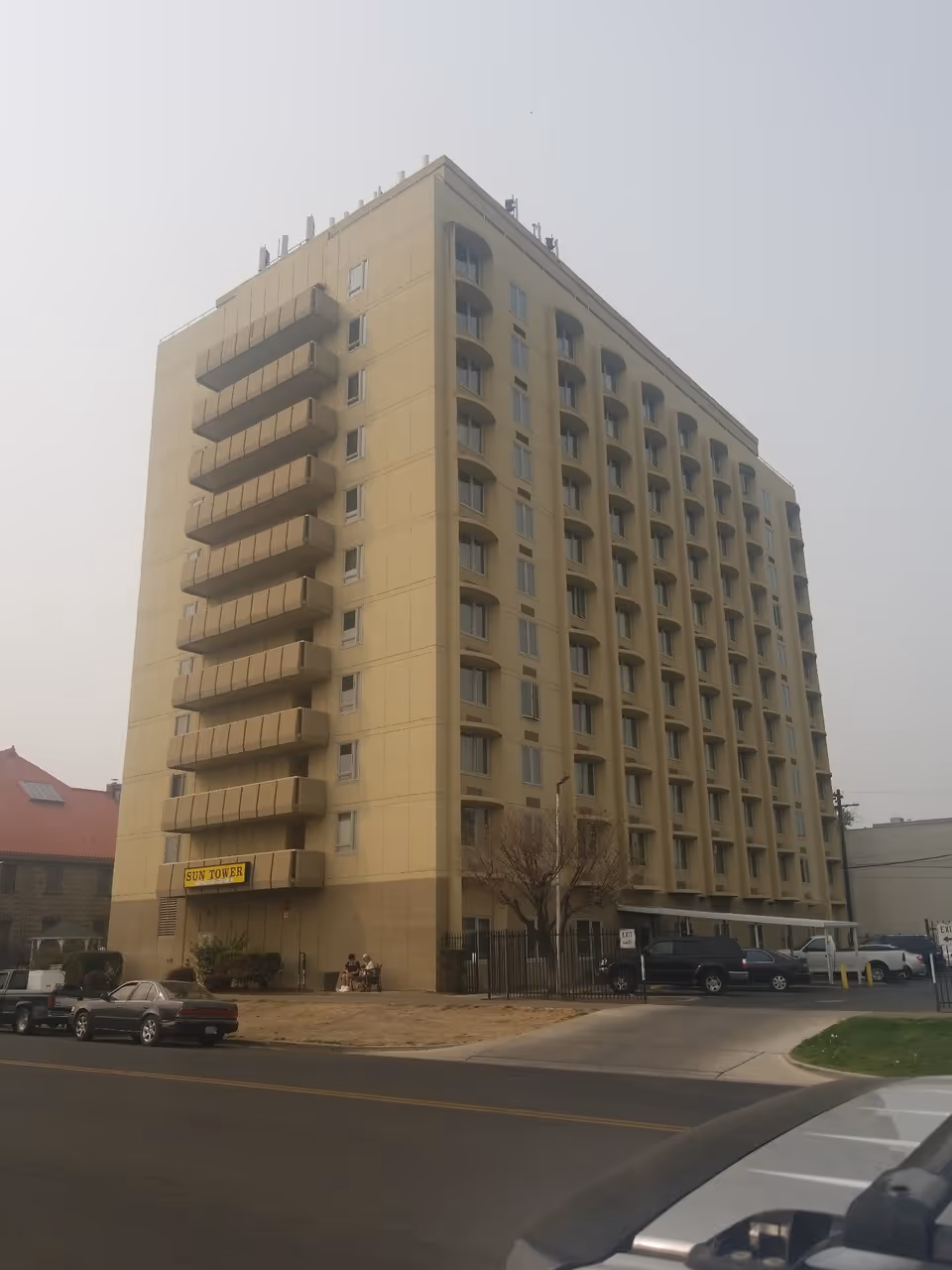 A multi-story beige building with balconies on one side and many windows on the other, identified as Sun Tower Retirement Home. There are several parked cars in front of the building and a few people sitting near the entrance. The sky is overcast.