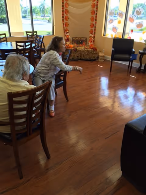 Two seated people in a communal senior living room with wooden floors, chairs, tables, and fall-themed decorations near the windows.