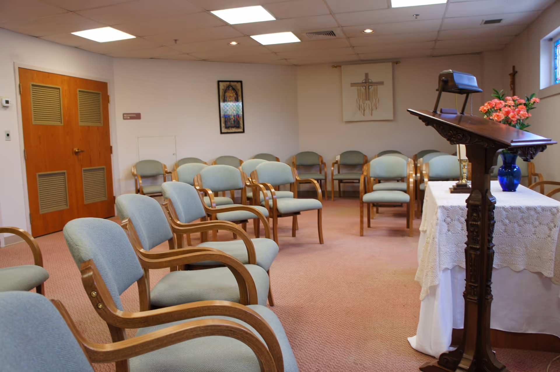 Small chapel-style meeting room with rows of wooden chairs facing a lectern and a table adorned with a lace cover and flowers.