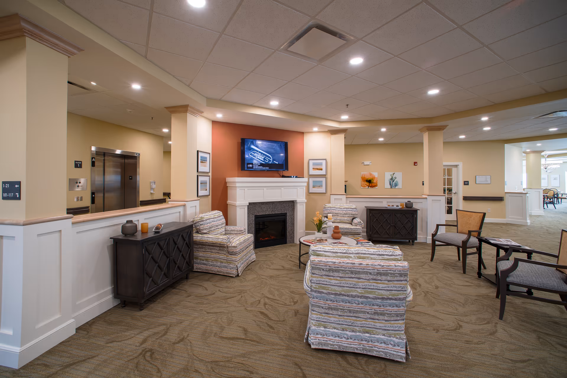 Comfortable senior living lounge with upholstered chairs arranged around a fireplace and wall-mounted TV.