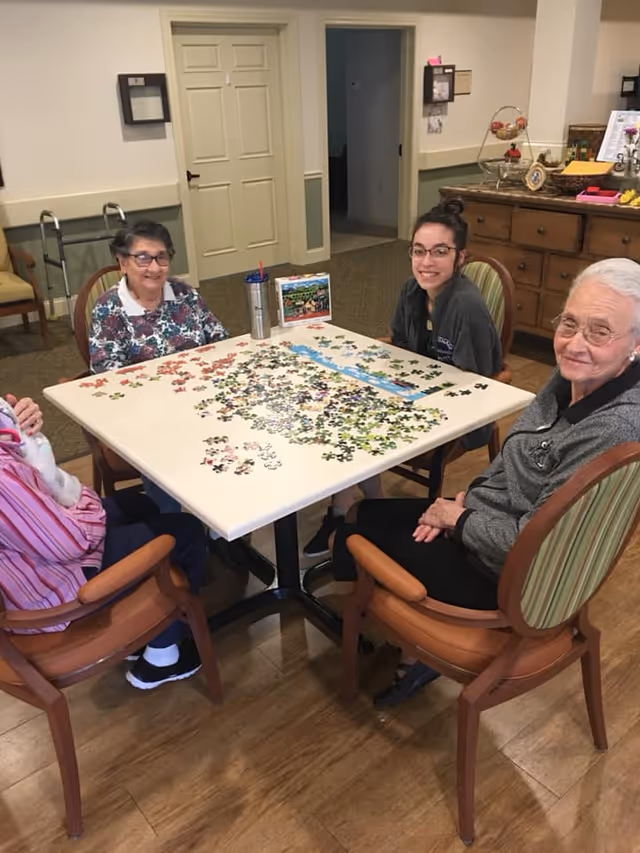 Four women sitting around a square table working on a jigsaw puzzle in a cozy room with wooden flooring and light-colored walls. There is a cabinet with drawers and some decorative items in the background.