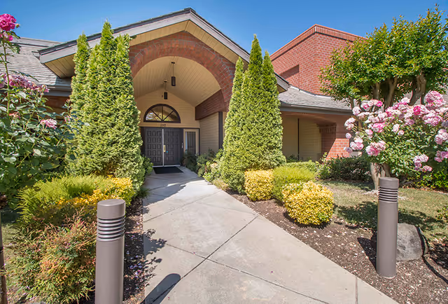 Entrance to a brick building with a paved walkway lined by shrubs and flowering bushes leading to double doors under a large arched porch.