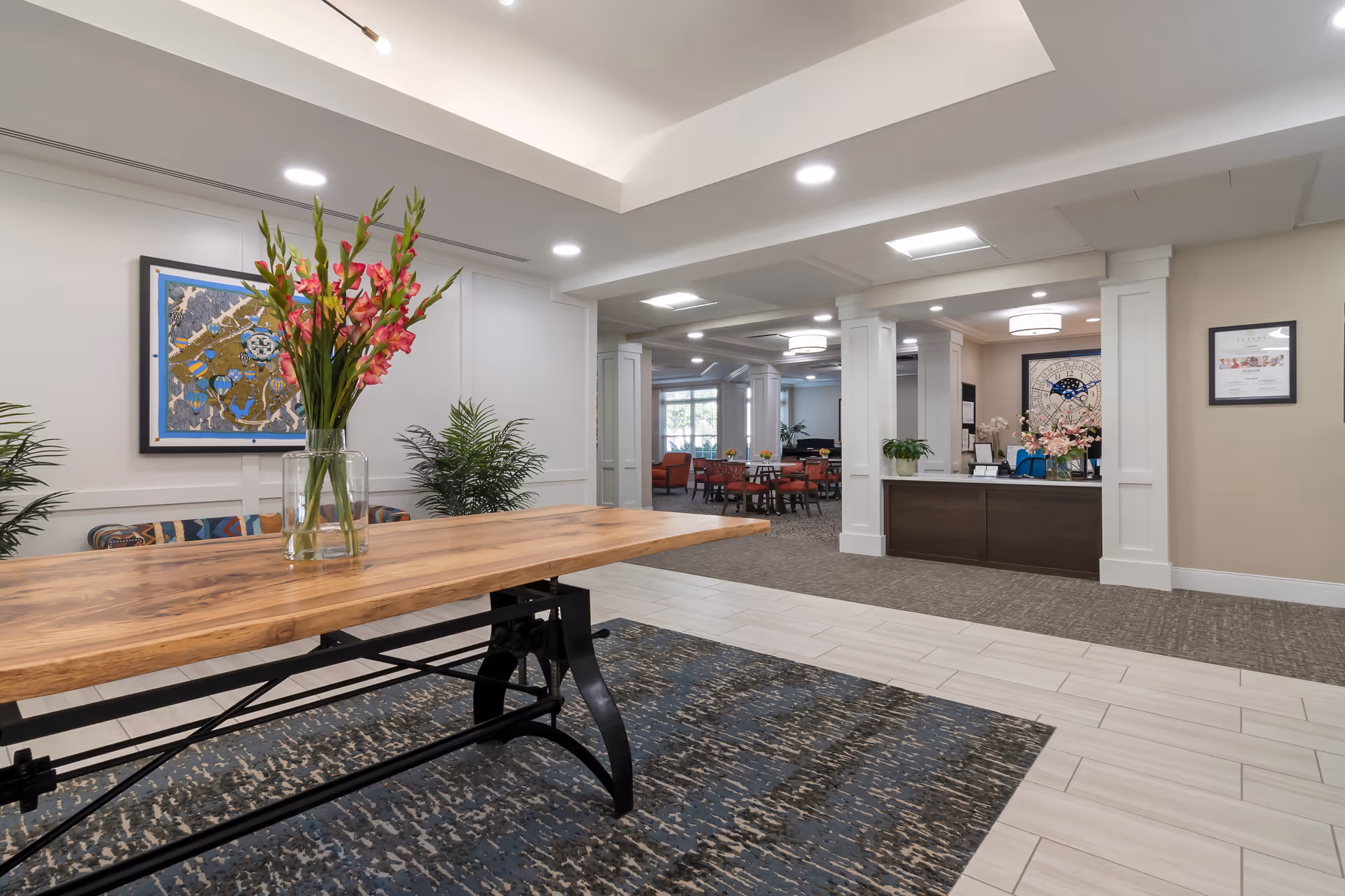 Interior view of a senior living facility lobby area with a wooden table holding a vase of pink flowers in the foreground. The background shows a reception desk, seating area with tables and chairs, decorative plants, framed artwork on the walls, and ceiling lights.