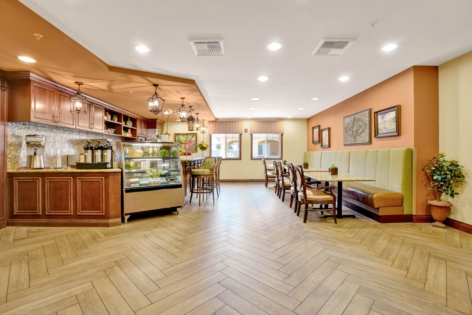 A bright and spacious dining area with a coffee bar on the left featuring wooden cabinets, a glass display case with pastries, and coffee machines. On the right, there are tables with chairs and a long green cushioned bench against the wall, decorated with framed artwork and a potted plant in the corner. The floor has a light wood herringbone pattern, and the ceiling has recessed lighting.