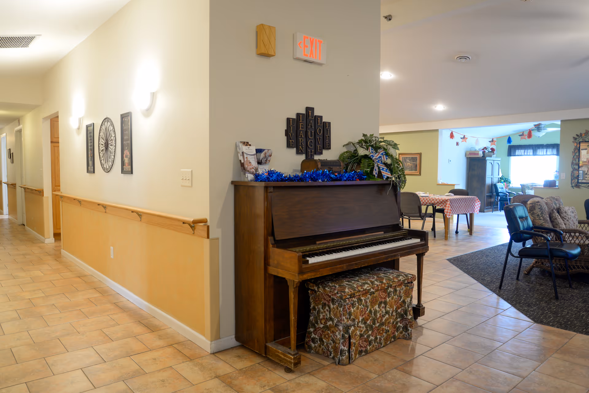 Interior view of a senior living facility hallway and common area. The hallway has tiled floors, light-colored walls with wooden handrails, and framed wall decorations. A wooden upright piano with a floral cushioned bench is placed at the corner near an exit sign. The common area features tables with checkered tablecloths, chairs, and a seating area with armchairs and a carpeted floor. There are some plants and decorative items on the piano and walls.