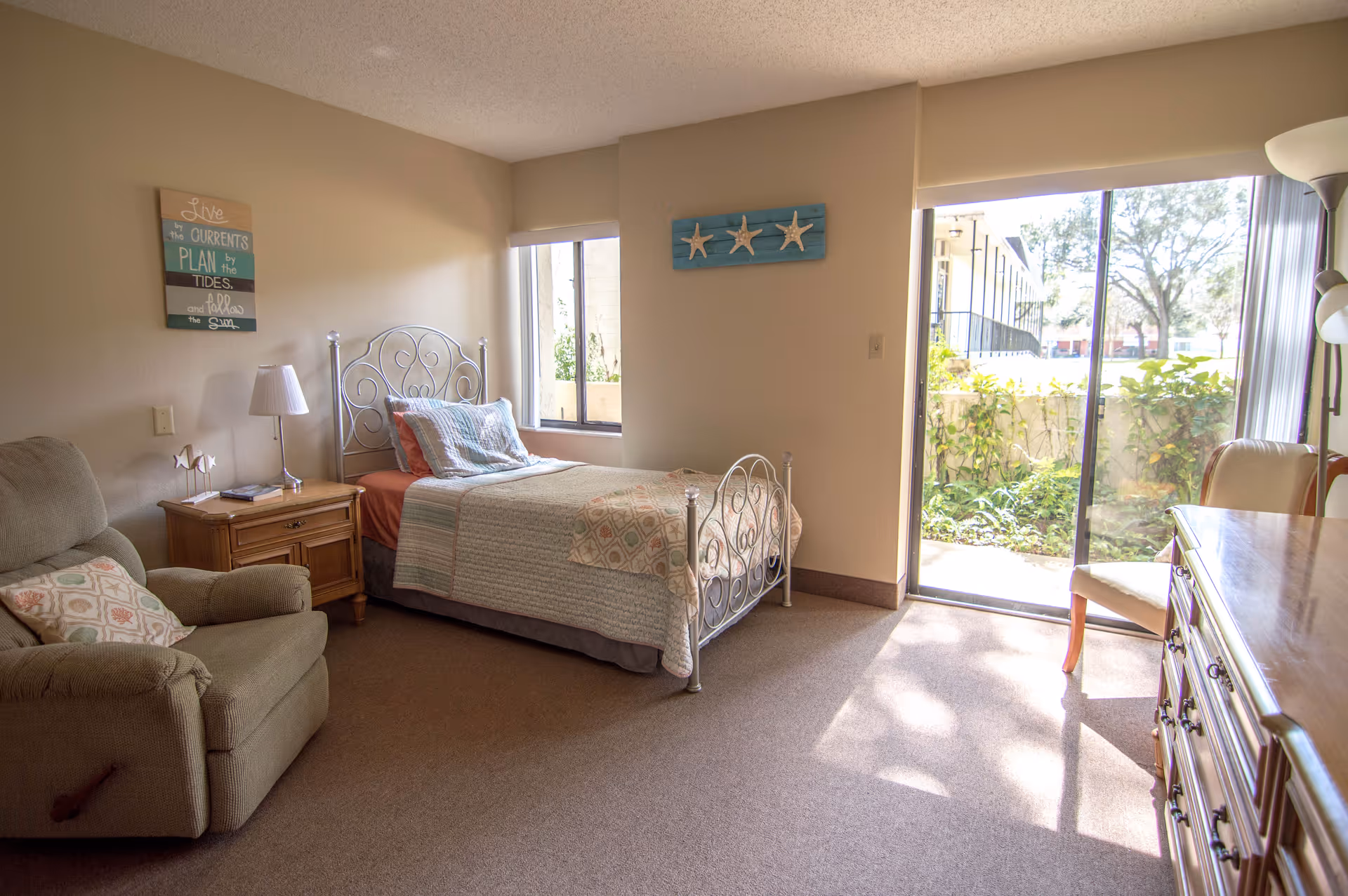 A cozy bedroom in a senior living facility featuring a single bed with a decorative metal headboard and footboard, a wooden nightstand with a lamp, a comfortable armchair with a patterned pillow, a dresser, and a sliding glass door leading to a small outdoor garden area. The room is decorated with wall art including a sign with an inspirational quote and a piece with three starfish.