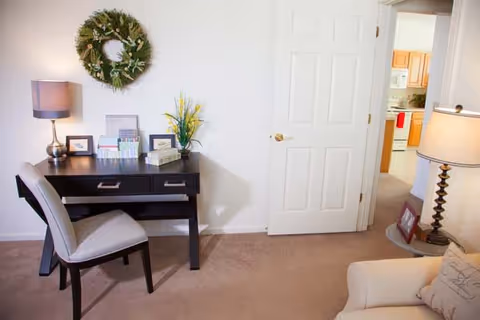 A cozy interior room with a black desk and a gray upholstered chair on a beige carpet. On the desk are a lamp, framed pictures, brochures, and a small plant. A green wreath hangs on the white wall above the desk. To the right, an open door reveals a kitchen area with wooden cabinets and white appliances. A beige armchair and a floor lamp with a beige shade are partially visible in the foreground.