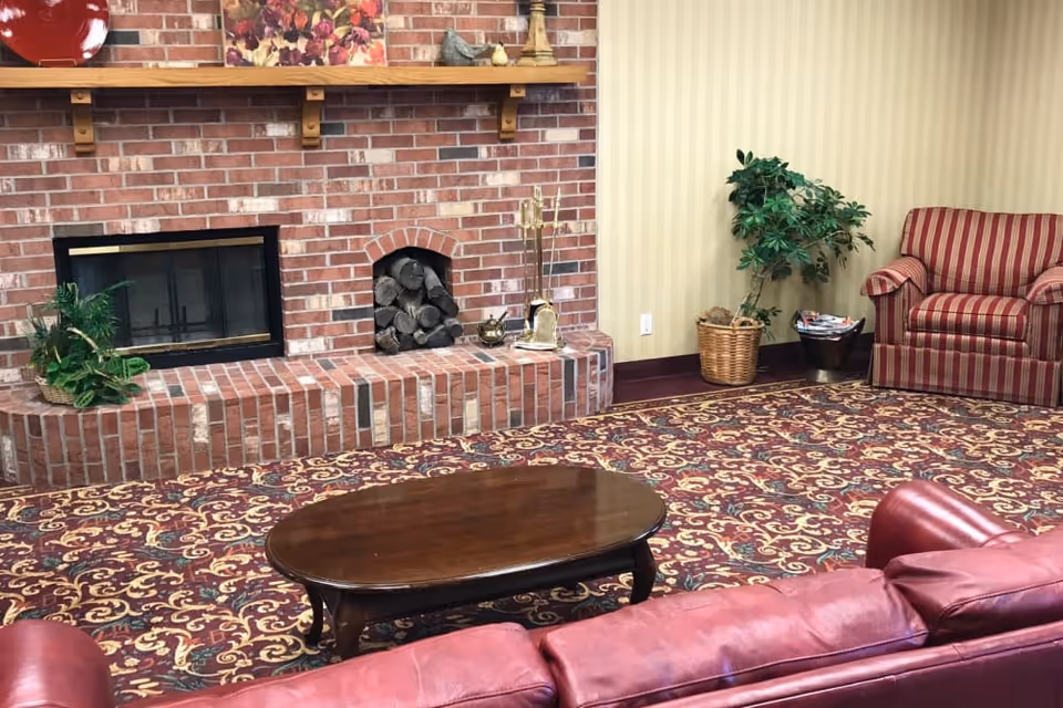A cozy living room with a red leather sofa and a red striped armchair. There is a wooden coffee table in front of the sofa. The room features a brick fireplace with firewood stored in a small arched niche and decorative items on the mantel. A potted plant is placed beside the armchair, and the floor is covered with a patterned carpet.