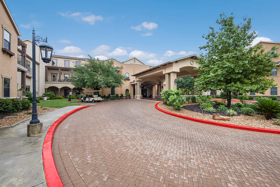 Entrance driveway of Villa de San Antonio senior living facility with a covered drop-off area, surrounded by landscaped greenery and multi-story beige buildings under a blue sky with some clouds.