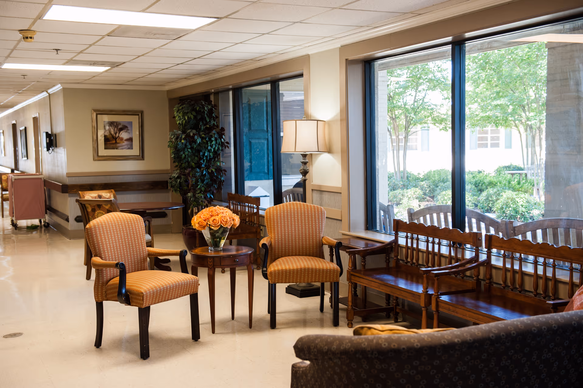 Sunlit senior living common area with upholstered chairs and a small table holding a vase of orange roses beside large windows and wooden benches.