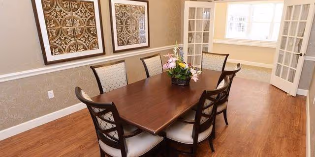 A dining room with a rectangular wooden table surrounded by six chairs, a floral centerpiece on the table, two framed decorative artworks on the wall, and French doors leading to a bright adjacent room with large windows.
