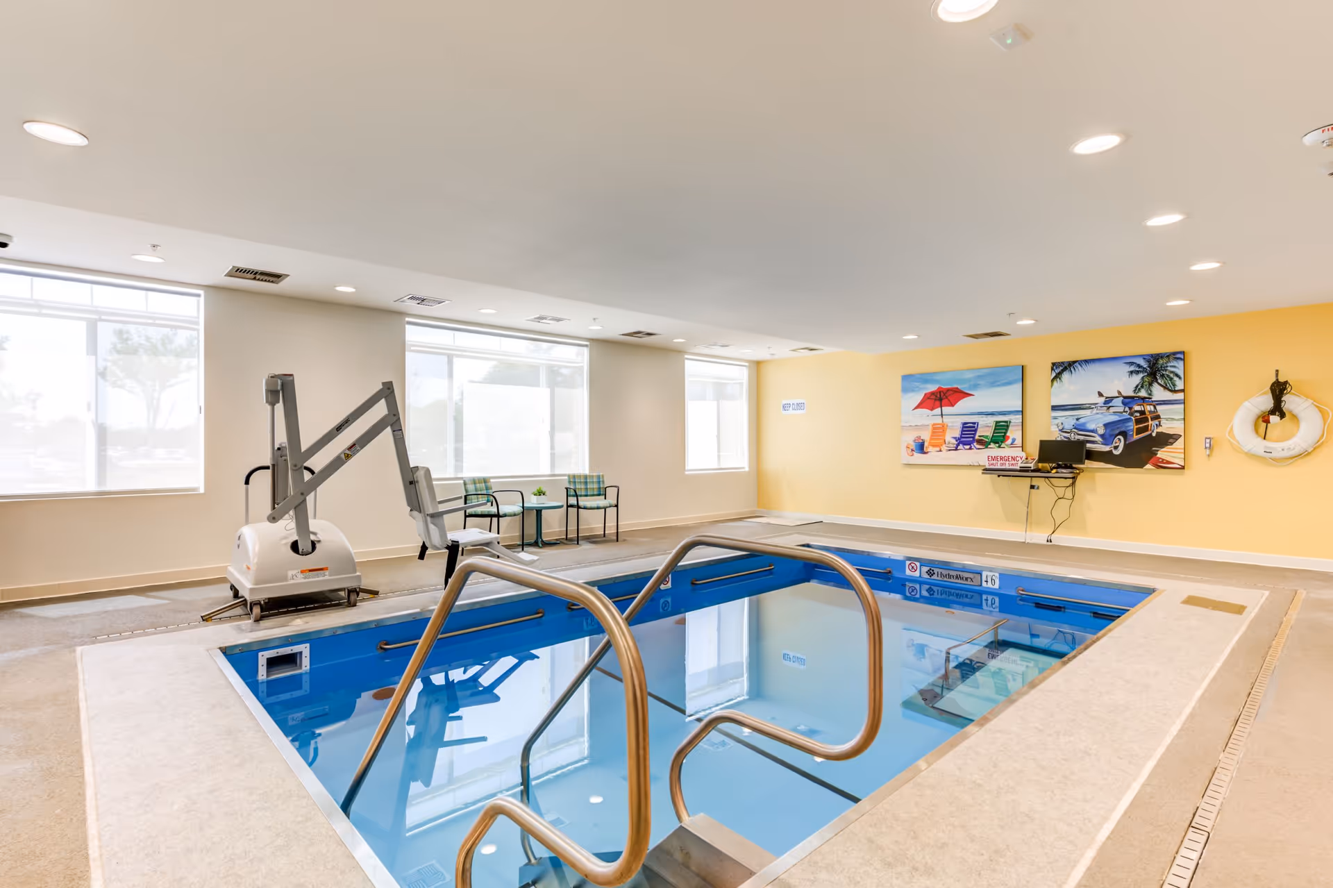 Indoor therapy pool with stainless handrails, a pool lift, seating and colorful wall art in a bright senior living facility room.