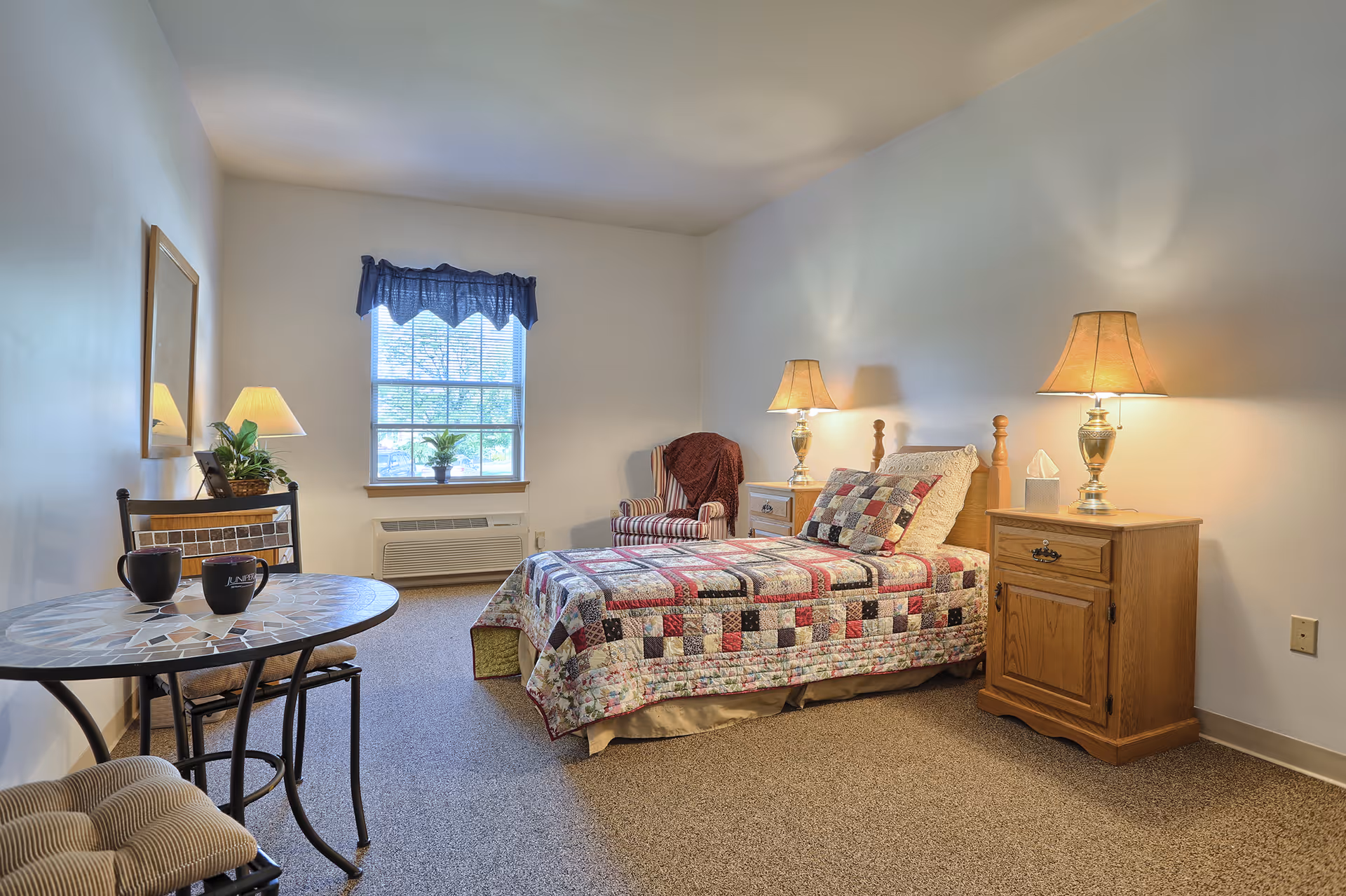 A cozy bedroom in a senior living facility featuring a single bed with a colorful patchwork quilt, two wooden nightstands with lamps, a striped armchair with a blanket, a window with a blue valance, and a small round table with two chairs and two black mugs.