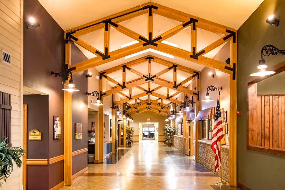 A well-lit hallway in a senior living facility with wooden beams and trusses on the ceiling, wall-mounted lights, an American flag on the right side, and plants placed along the corridor. The hallway has a polished floor and doorways leading to other rooms on both sides.