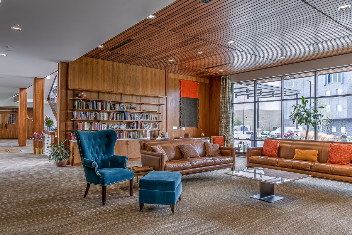 A modern and spacious living room area with two brown leather sofas adorned with colorful cushions, a teal upholstered armchair with matching ottoman, a glass coffee table, and a wooden bookshelf filled with books. Large floor-to-ceiling windows provide natural light and a view of the outdoor area. The ceiling and some walls are covered with wood paneling, and there are plants placed around the room.
