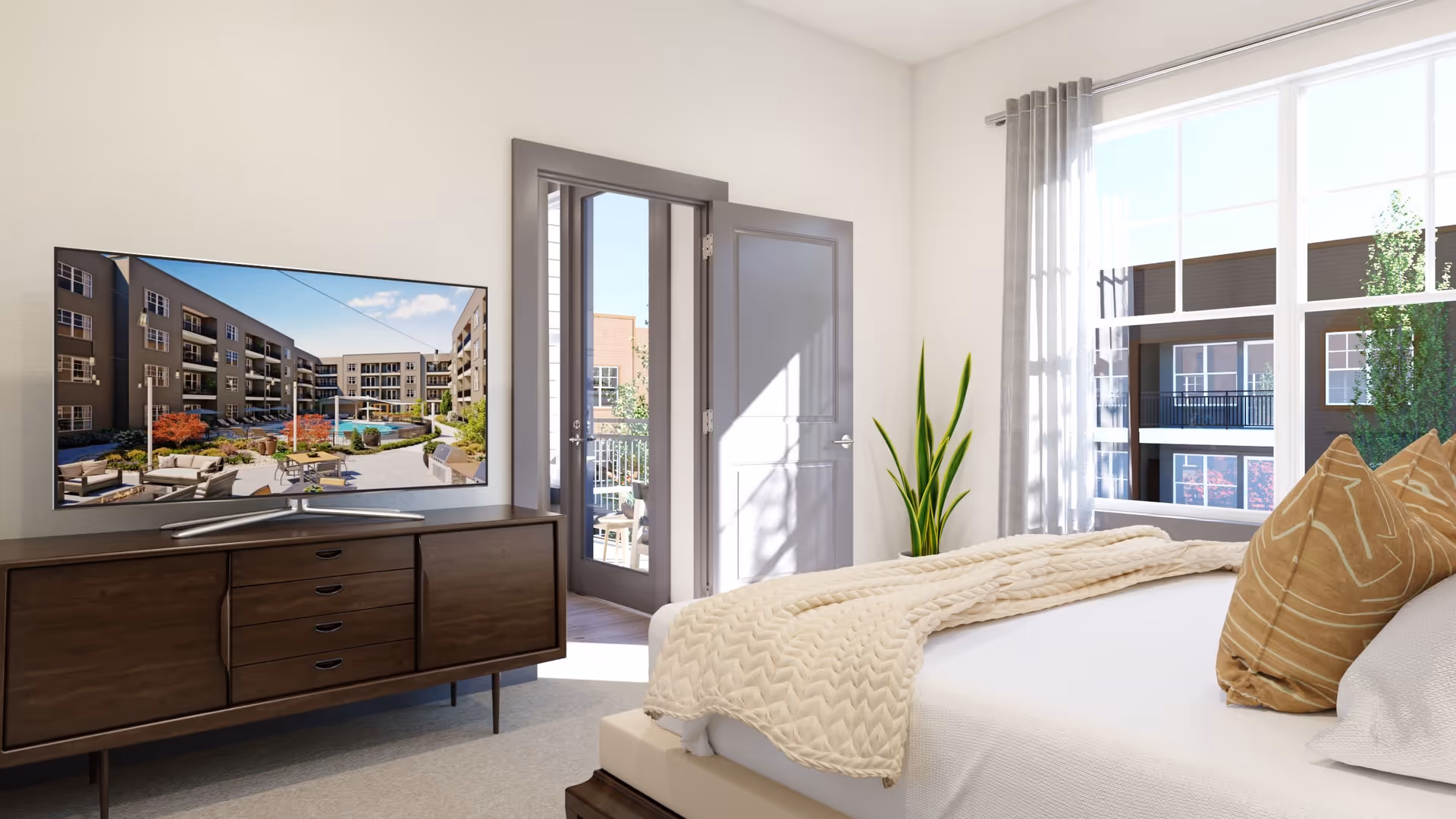 A bright bedroom with a neatly made bed featuring beige pillows and a cream knitted blanket. A wooden dresser with a flat-screen TV displaying an outdoor courtyard with seating and a pool is against the wall. A door next to the dresser opens to a balcony, and a large window with gray curtains lets in natural light, showing an exterior view of a building and trees.
