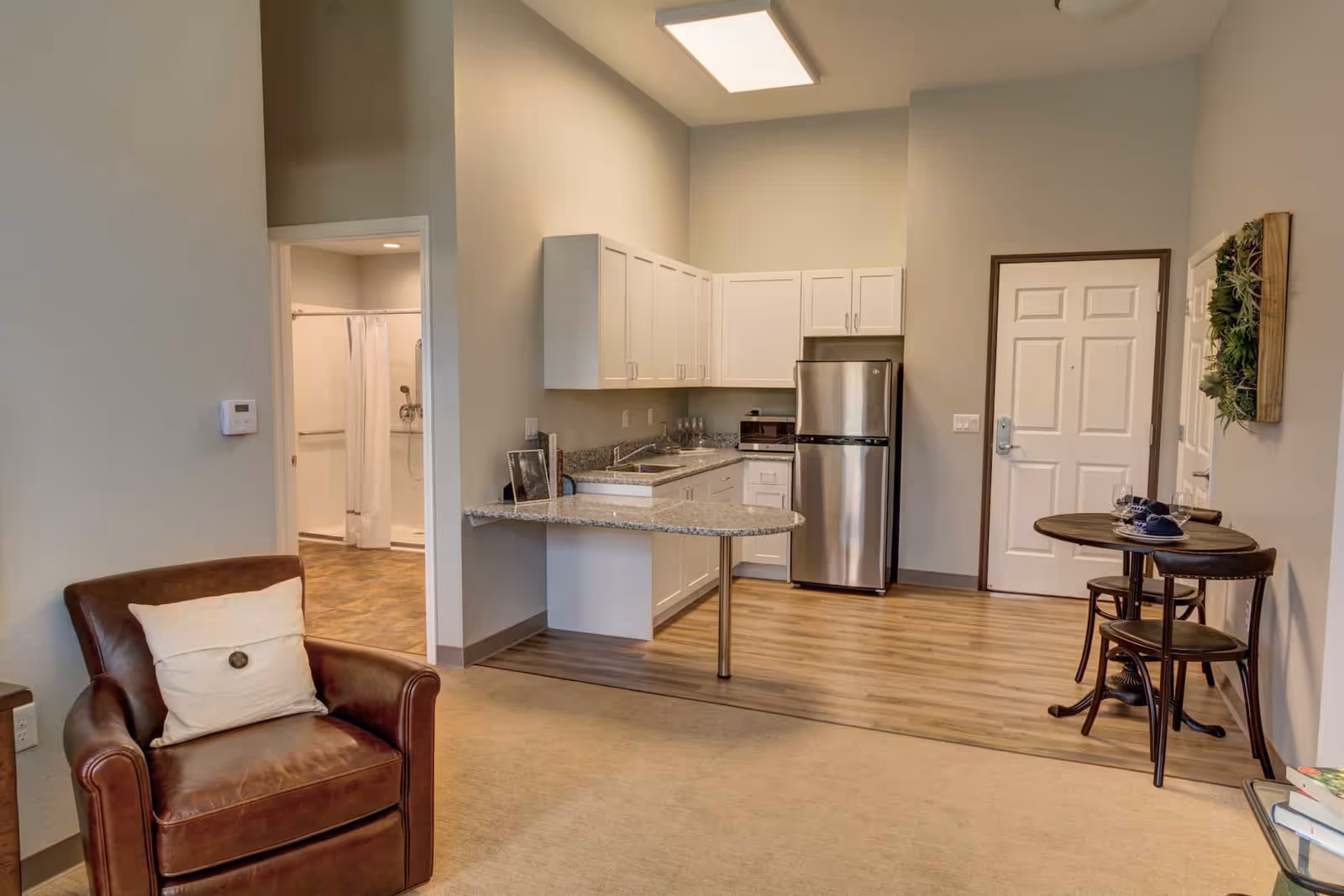Interior view of a senior living facility apartment showing a small kitchen area with white cabinets, a stainless steel refrigerator, and a granite countertop with a breakfast bar. To the right, there is a small round dining table with two chairs. On the left side, there is a brown leather armchair with a white pillow. In the background, an open door reveals a bathroom with a shower and grab bars. The flooring transitions from carpet in the living area to wood in the kitchen and bathroom areas.