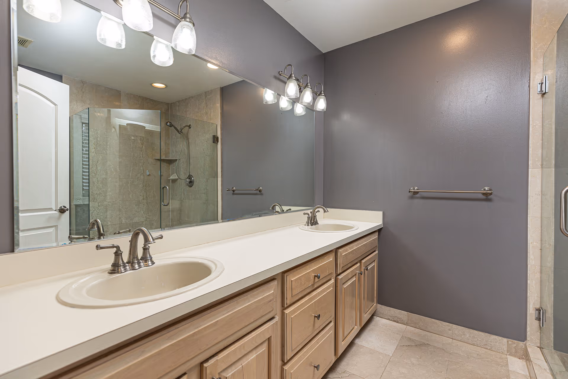 A modern bathroom with a double sink vanity featuring light wood cabinets and white countertops. Above the sinks is a large mirror with three light fixtures mounted on the wall. The walls are painted gray, and there is a glass-enclosed shower with beige tiles visible in the background. A towel bar is mounted on the wall next to the vanity.