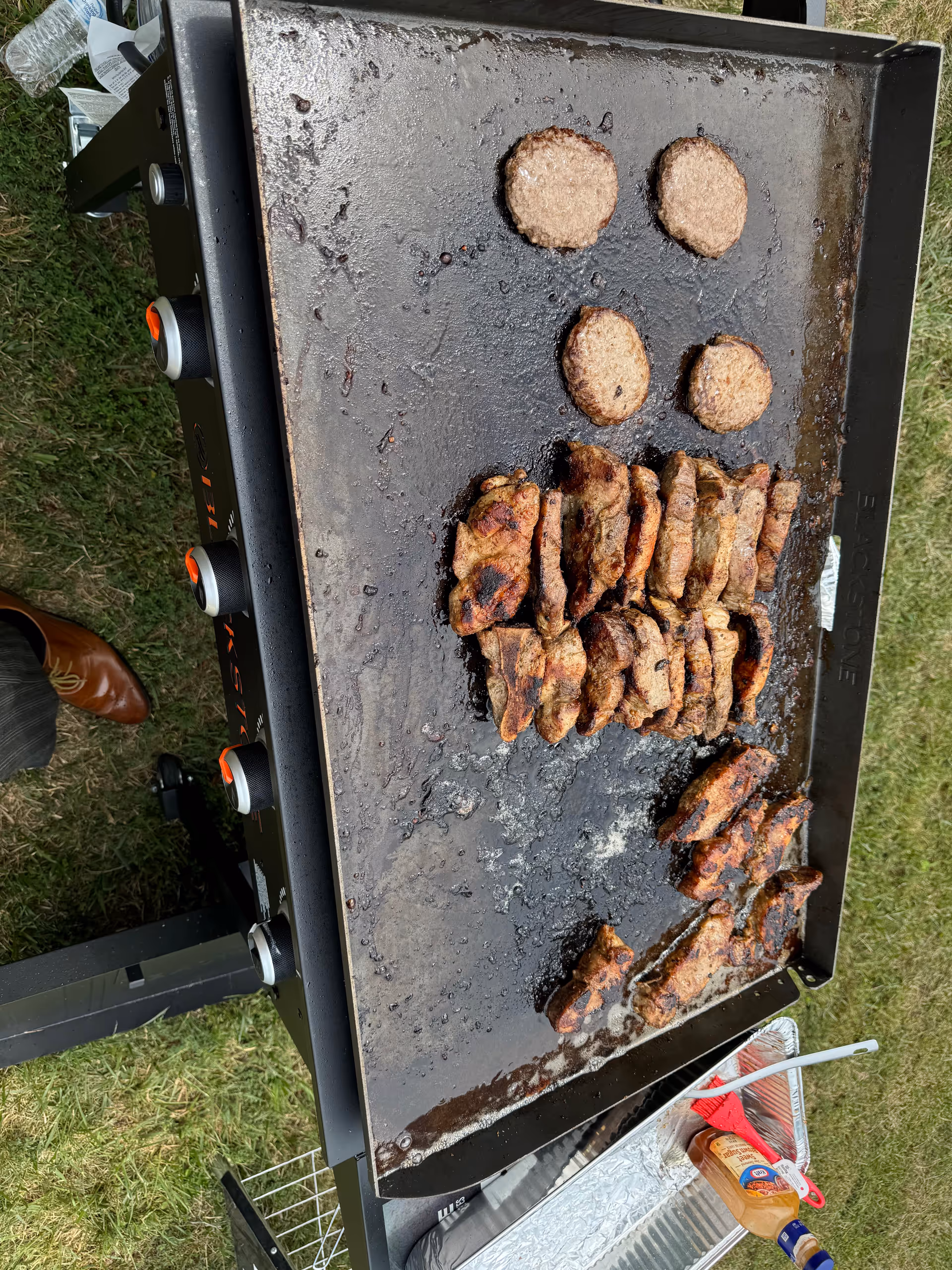 A large outdoor griddle with several cooked hamburger patties and grilled chicken pieces on it. The griddle is set up on a grassy area with a person's brown shoe visible on the left side. There is a foil tray with a bottle of barbecue sauce and a red basting brush nearby.