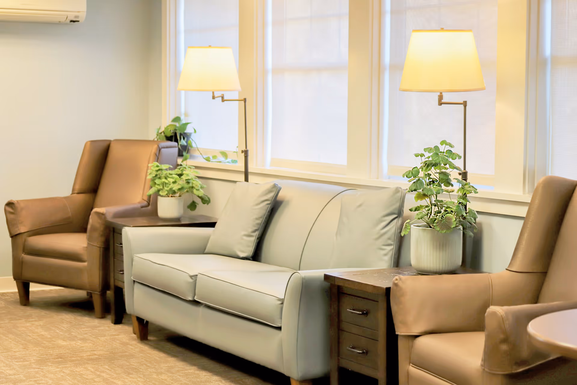 Comfortable seating area with a sofa, armchairs, side tables, lamps, and potted plants in a bright common room.