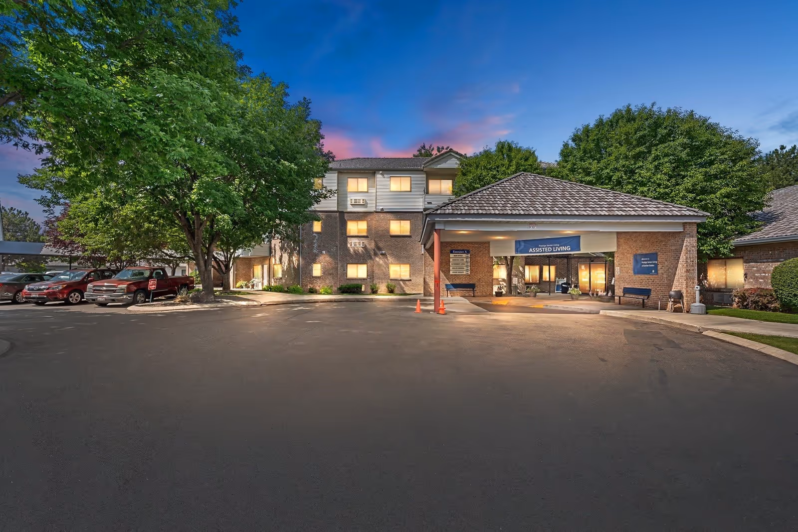 Exterior view of Karcher Senior Living facility at dusk with a parking area, several parked cars, large trees, and a covered entrance labeled 'Assisted Living'. The building has multiple windows lit from inside and a tiled roof.