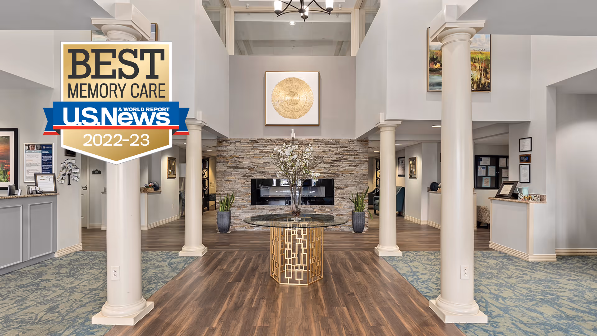 Spacious and elegant interior of a senior living facility lobby with tall white columns, a round glass table with a decorative vase in the center, wood flooring, and a stone accent wall with a modern fireplace. Artwork is displayed on the walls and there is a reception desk on the left side.