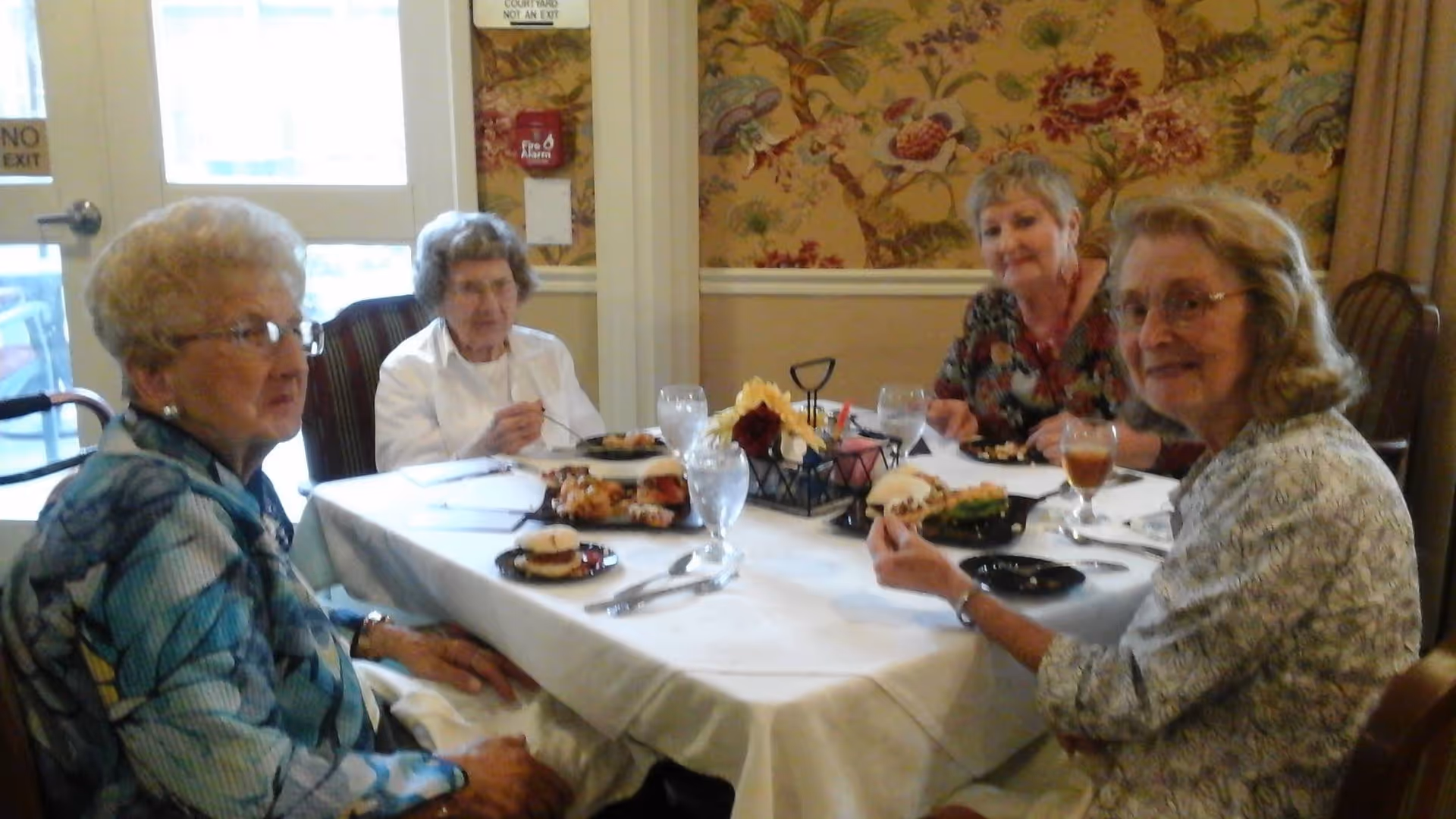 Four elderly women seated around a table eating a meal in a decorated dining room.