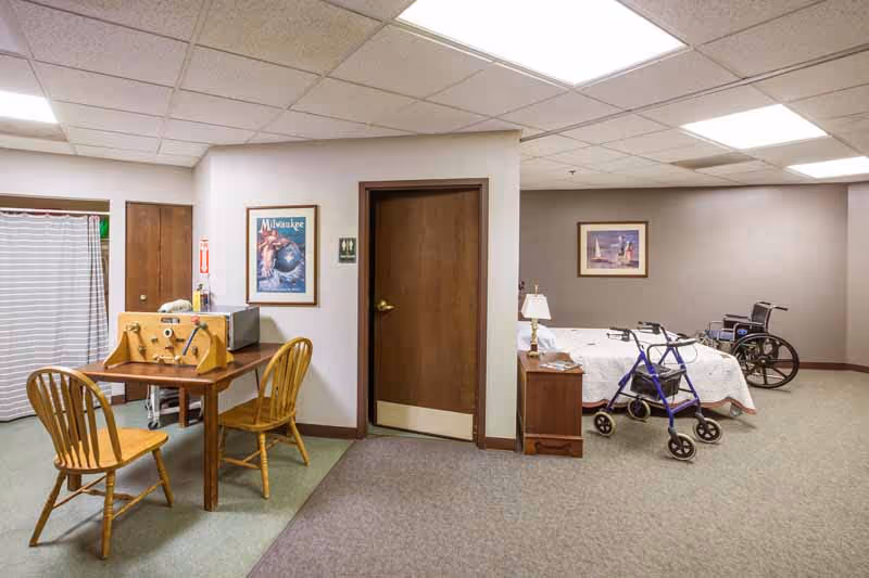 Interior view of a senior living facility room with a small wooden table and two chairs on the left, a closed wooden door in the center, and a hospital-style bed with a walker and wheelchair on the right. The walls have framed pictures, and the ceiling has fluorescent lighting.