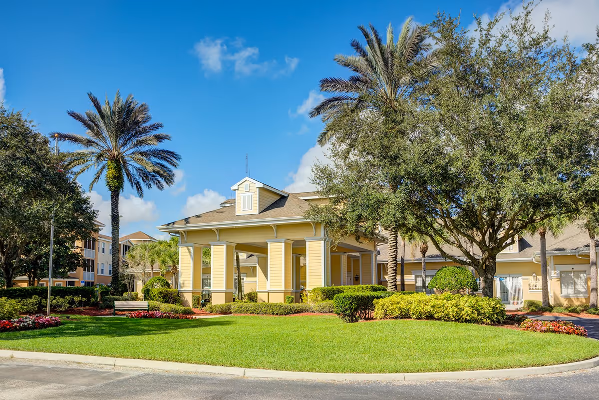 Exterior view of a senior living facility with a yellow covered entrance surrounded by green lawns, palm trees, and other landscaping under a blue sky with some clouds.