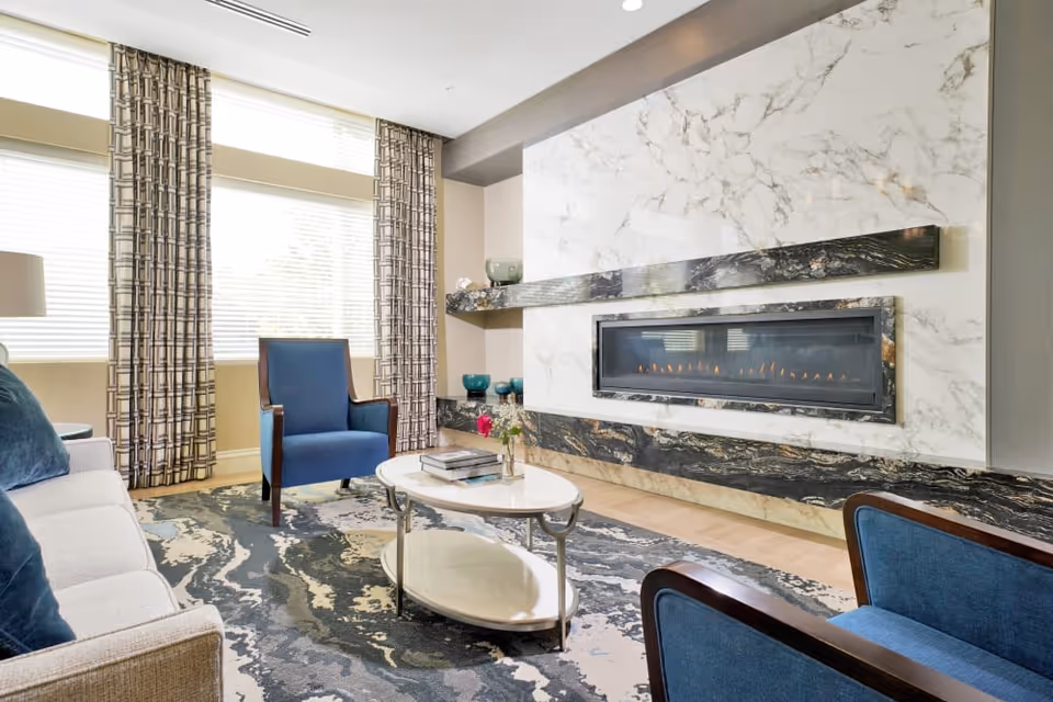 A modern living room with a white sofa, two blue armchairs, a white oval coffee table with books and a small vase of flowers, a patterned area rug, large windows with patterned curtains, and a sleek gas fireplace set in a marble and dark stone wall.